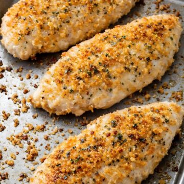 Three baked parmesan chicken breasts on a metal sheet pan, topped with a golden crispy breadcrumb and parmesan crust with herbs and black pepper. Juicy white chicken with toasted crumbs scattered around the pan.