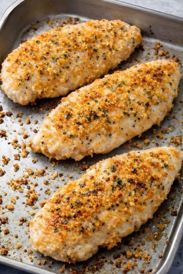 Three baked parmesan chicken breasts on a metal sheet pan, topped with a golden crispy breadcrumb and parmesan crust with herbs and black pepper. Juicy white chicken with toasted crumbs scattered around the pan.
