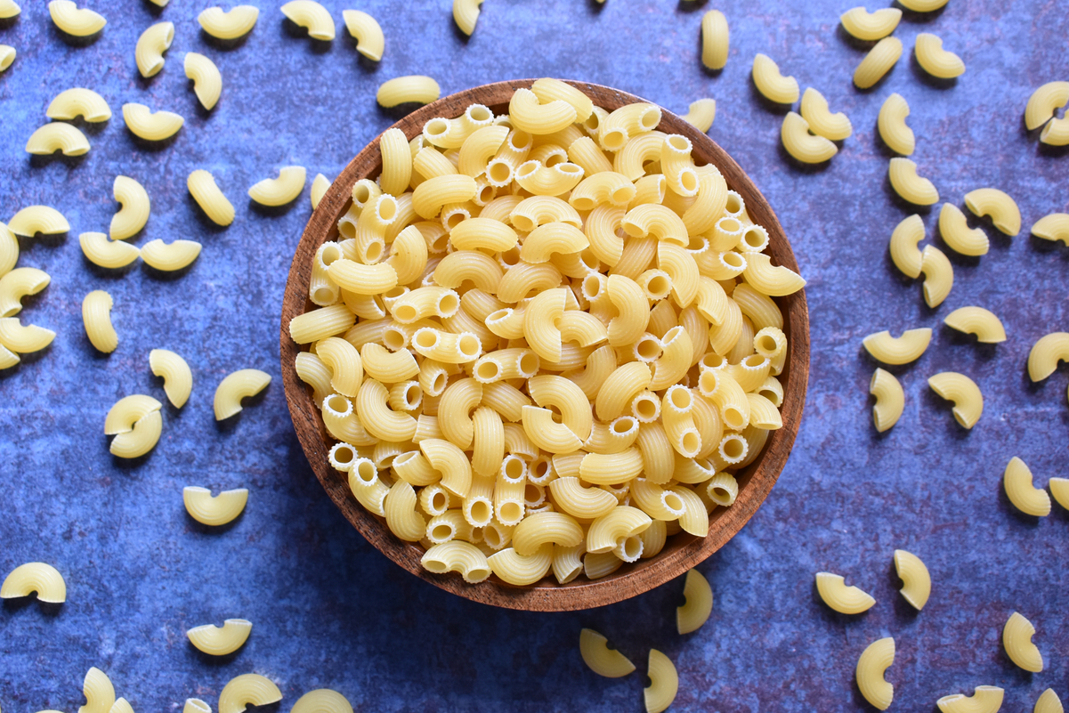 Dry macaroni in a brown bowl on a blue surface, surrounded by spilled macaroni.