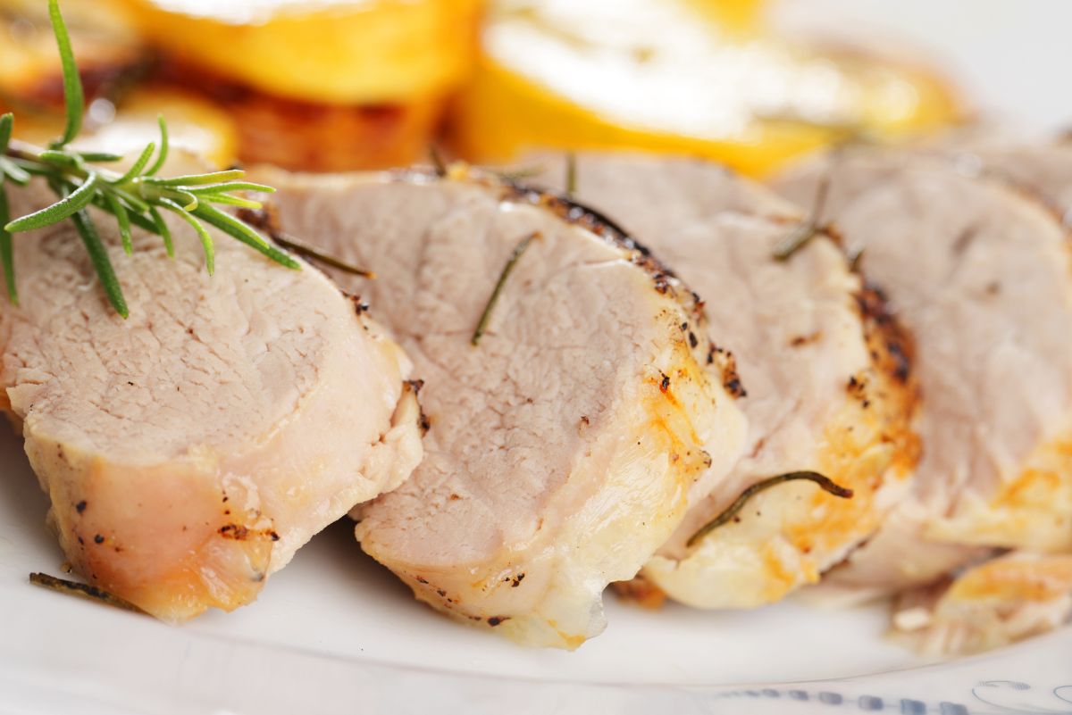 Sliced roasted pork tenderloin on a white plate, lightly browned on the outside and juicy inside, seasoned with herbs and garnished with fresh rosemary. Golden roasted potatoes are blurred in the background, highlighting a simple, high protein pork dinner.