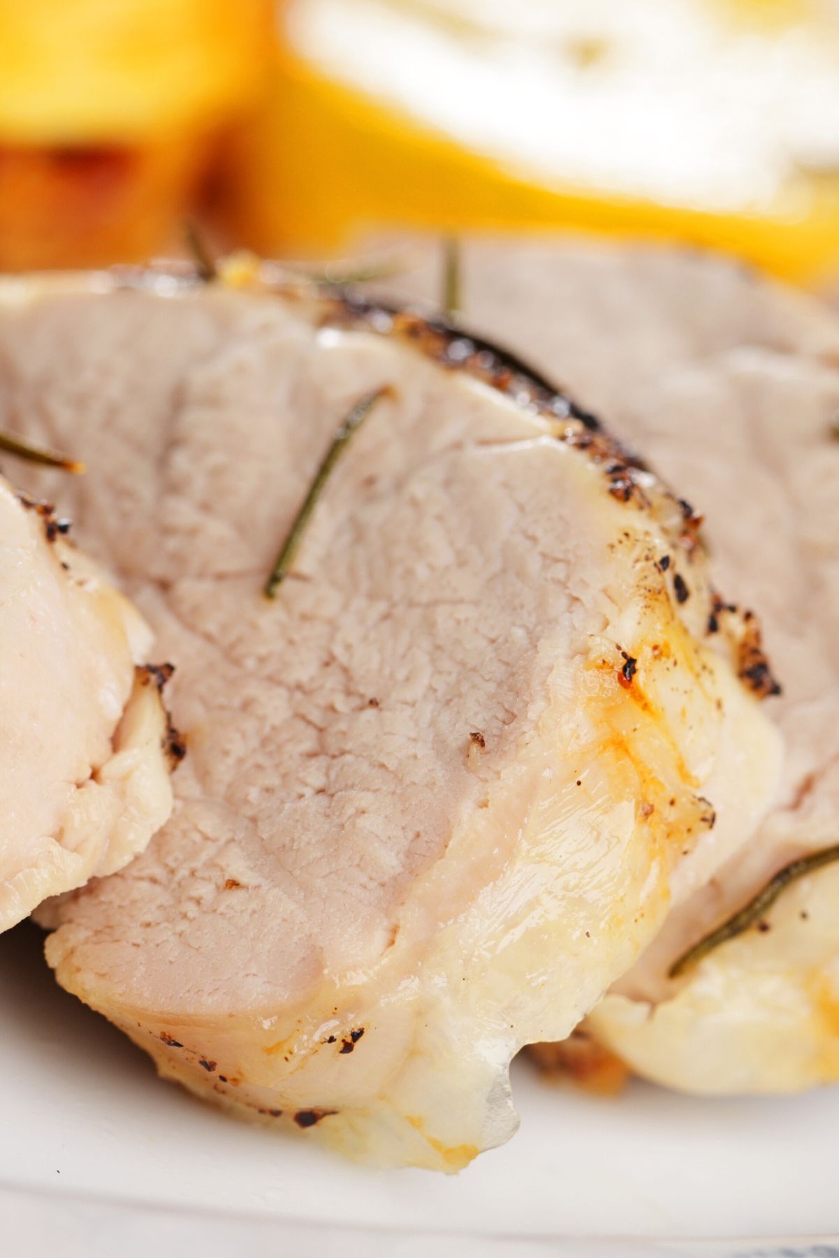 Close-up of sliced roasted pork tenderloin with a lightly browned edge and juicy interior, seasoned with herbs and small rosemary needles. The pork sits on a white plate, with golden roasted potatoes softly blurred in the background, highlighting a simple high protein pork dinner.