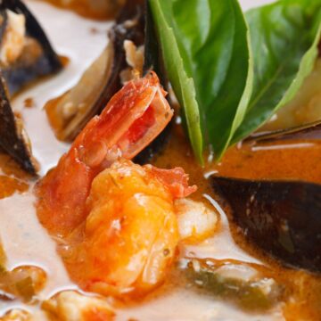 Close-up of San Francisco style cioppino seafood stew with shrimp, mussels, and white fish in a rich tomato-based broth. Fresh basil leaves sit on top, with visible vegetables and seafood in a creamy, savory sauce, highlighting a high protein seafood stew.