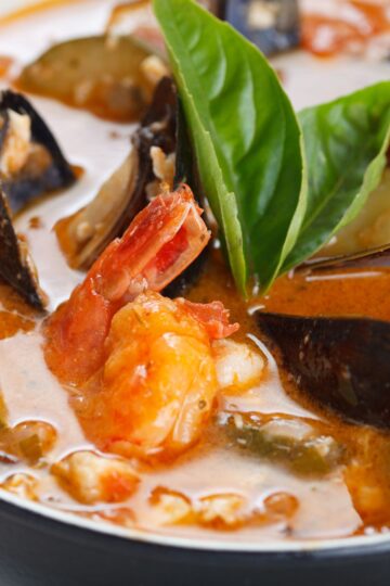 Close-up of San Francisco style cioppino seafood stew with shrimp, mussels, and white fish in a rich tomato-based broth. Fresh basil leaves sit on top, with visible vegetables and seafood in a creamy, savory sauce, highlighting a high protein seafood stew.