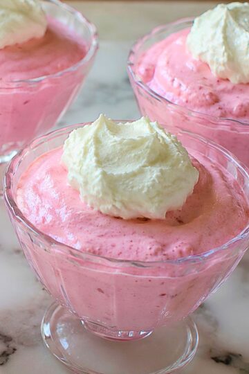 Cherry yogurt Jello fluff served in clear glass dessert cups with whipped topping, showing a creamy pink no-bake dessert on a marble countertop.