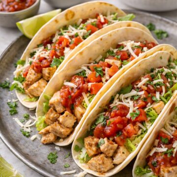 Four soft chicken tacos on a round plate, filled with diced seasoned chicken, lettuce, salsa, chopped tomatoes, cilantro, and shredded cheese. Lime wedges, salsa, and extra toppings sit in the background on a light countertop.