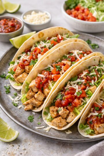 Four soft chicken tacos on a round plate, filled with diced seasoned chicken, lettuce, salsa, chopped tomatoes, cilantro, and shredded cheese. Lime wedges, salsa, and extra toppings sit in the background on a light countertop.