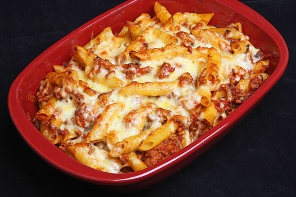 Baked ziti in a red casserole dish. A solid black background.