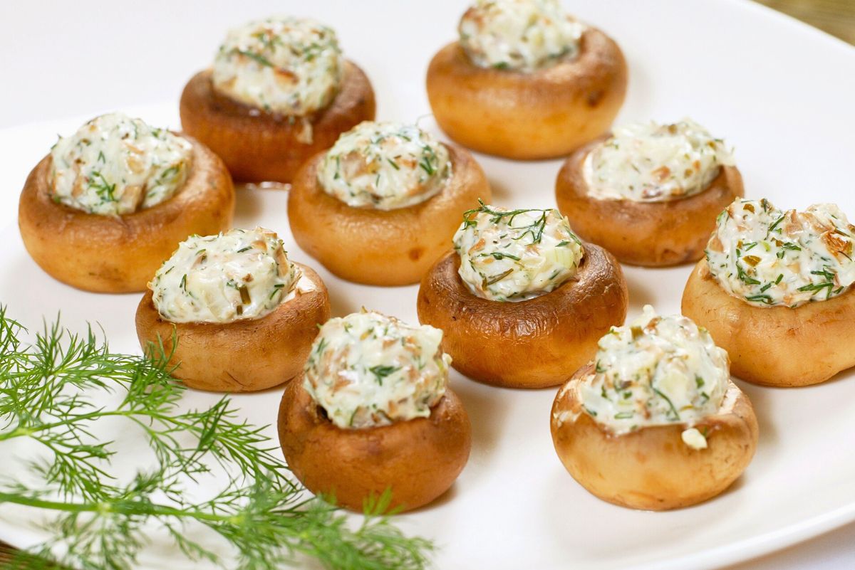 Baked artichoke stuffed mushrooms arranged on a white plate, each mushroom cap filled with a creamy artichoke and herb mixture. Golden brown mushrooms with visible flecks of dill in the filling, garnished with fresh dill sprigs on the side in bright, clean food photography.
