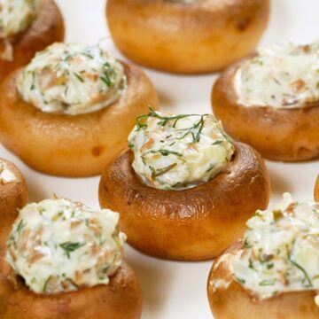 Artichoke stuffed mushrooms baked until golden brown, filled with a creamy artichoke and herb mixture. Mushrooms are arranged on a white plate with visible flecks of fresh dill, showing a smooth, cheesy filling and tender mushroom caps in close-up food photography.