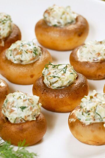 Artichoke stuffed mushrooms baked until golden brown, filled with a creamy artichoke and herb mixture. Mushrooms are arranged on a white plate with visible flecks of fresh dill, showing a smooth, cheesy filling and tender mushroom caps in close-up food photography.