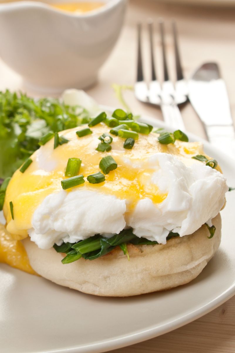 Closeup of eggs benedict on a white plate with flatware behind it.