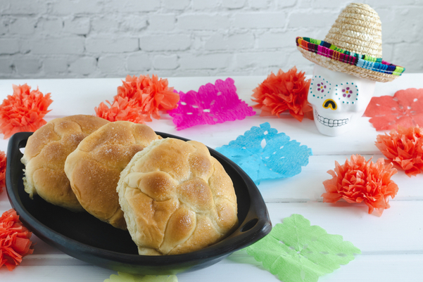 3 loaves of Weight Watchers Day of the Dead Bread (Pan de Muerto) on a black plate with a sugar skill in the background and colorful paper decorations around it.