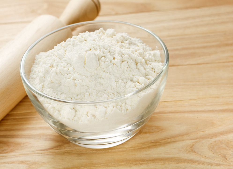 A glass bowl of flour on a wooden surface with a rolling pin next to it.