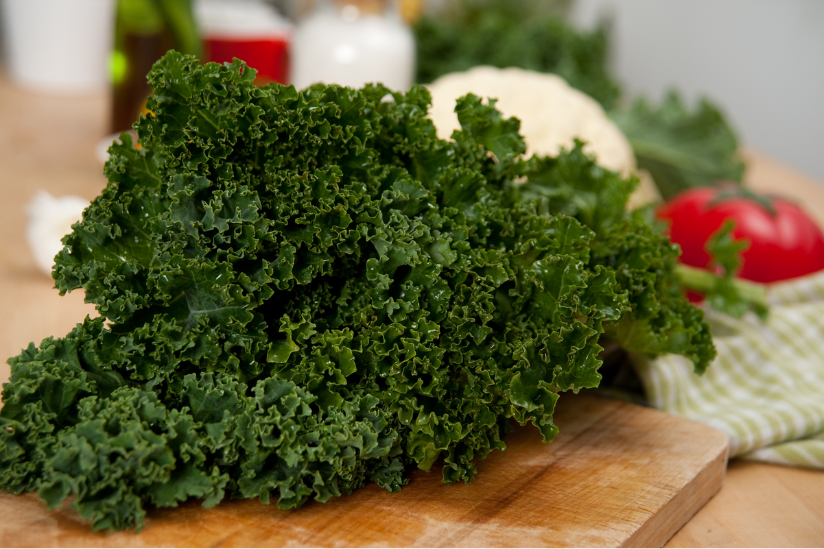 Fresh kale on a cutting board with out of focus vegetables in the background.
