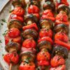 Grilled vegetable kabobs on a large plate with charred cremini mushrooms, cherry tomatoes, red bell pepper chunks, and red onion on skewers. The vegetables are glossy with oil and herbs, with light grill marks throughout. Perfect summer grilling recipe or healthy vegetable skewers.