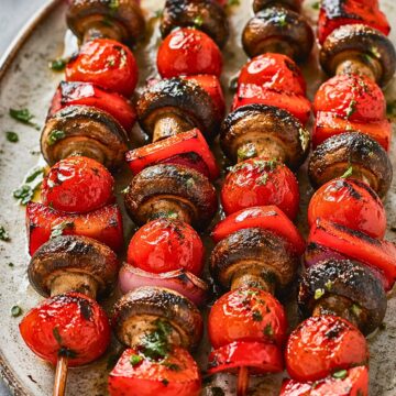 Grilled vegetable kabobs on a large plate with charred cremini mushrooms, cherry tomatoes, red bell pepper chunks, and red onion on skewers. The vegetables are glossy with oil and herbs, with light grill marks throughout. Perfect summer grilling recipe or healthy vegetable skewers.