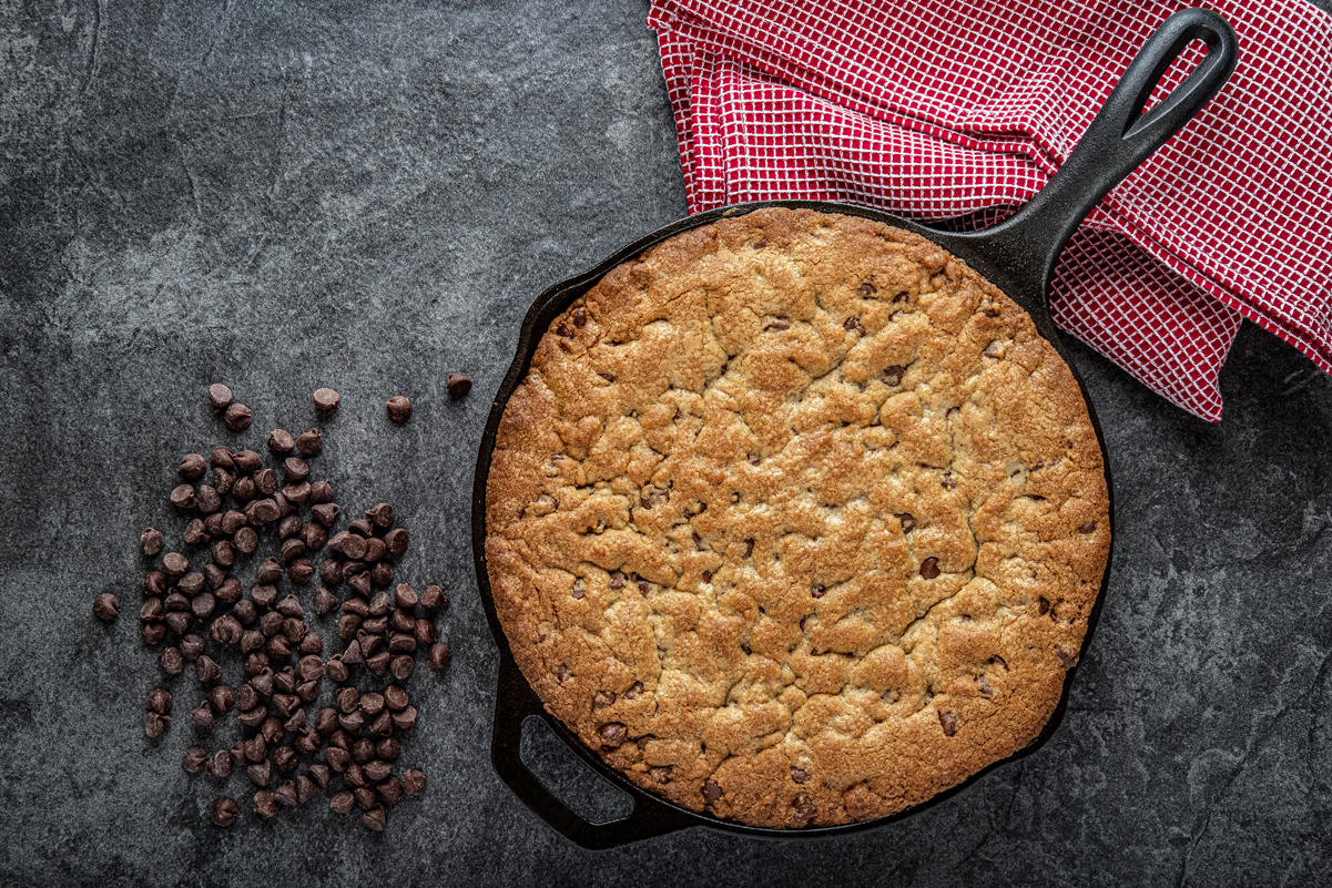 Grilled Skillet Chocolate Chip Peanut Cookie in a cast iron skillet on a slate surface with chocolate chips and a red and white napkin next to it.