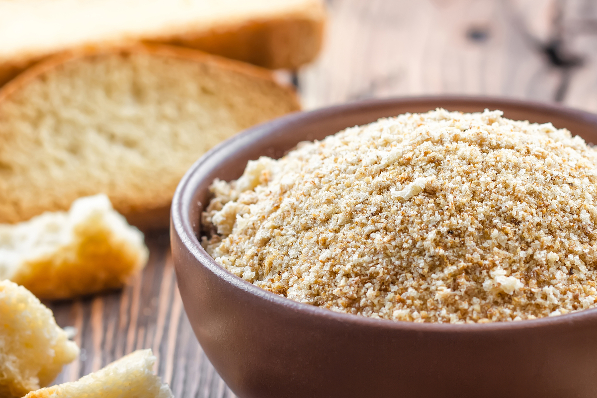 Breadcrumbs in a brown, wooden bowl with sliced bread behind it.