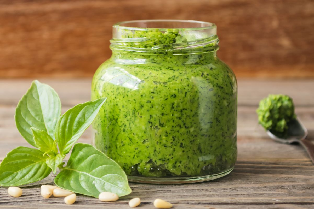 Pesto sauce in a small, clear glass jar with fresh basil leaves and pine nuts next to it. All sitting on a rustic wooden surface.