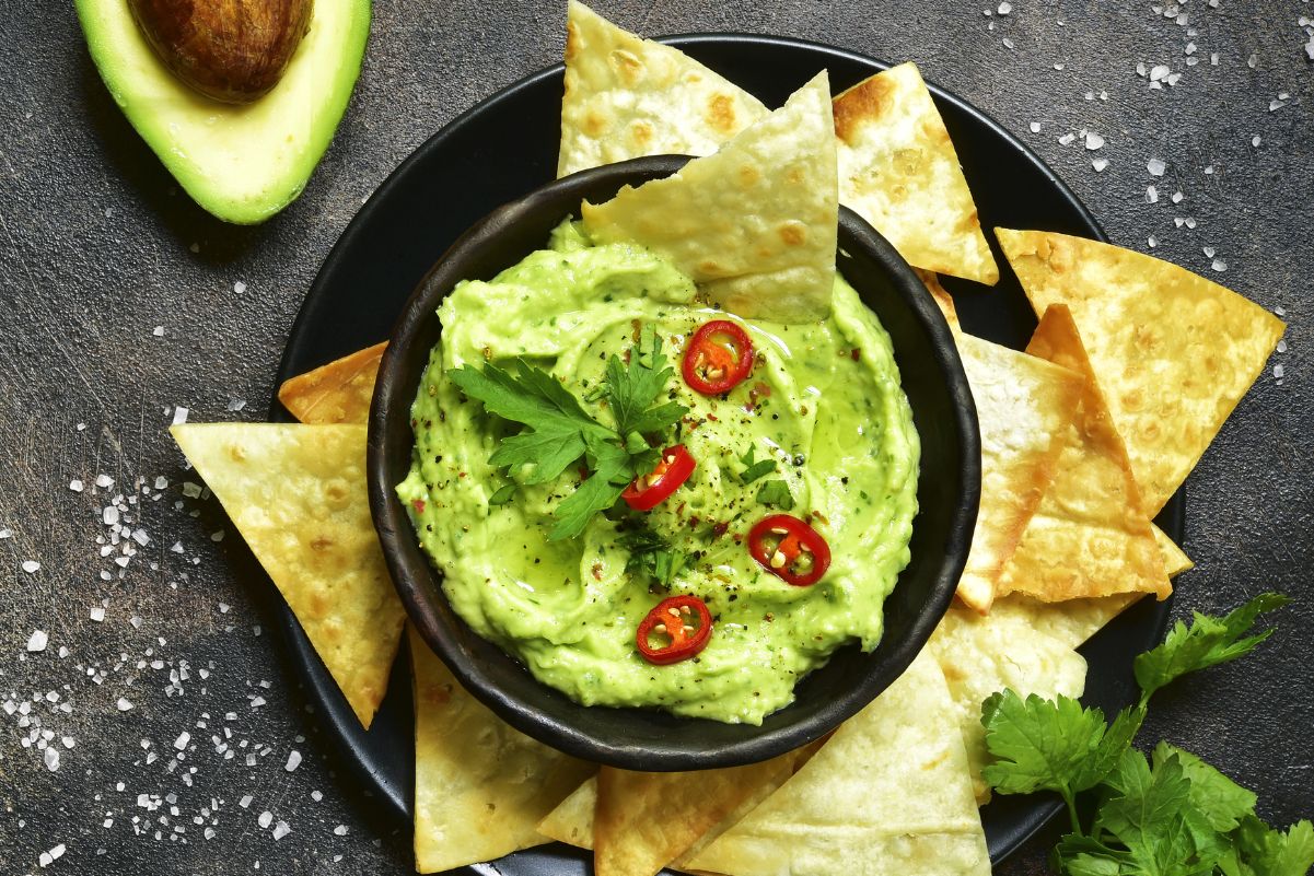 Overhead view of Simple Weight Watchers Whipped Guacamole in a black bowl on a black dish with tortilla chips. A fresh avocado in the background.