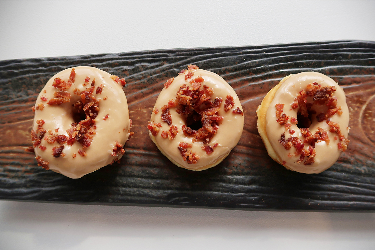 3 Maple Glazed Doughnuts with Bacon on a piece of rustic wood sitting on a white surface.
