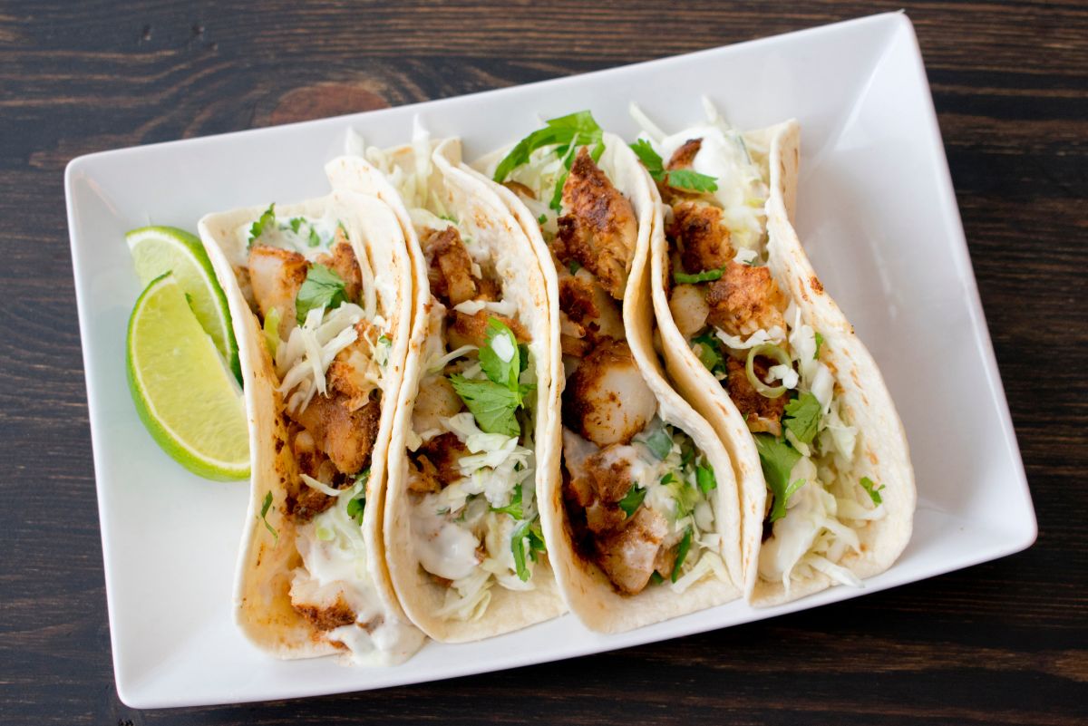 4 California Style Fish Tacos on a white plate sitting on a dark wooden surface.