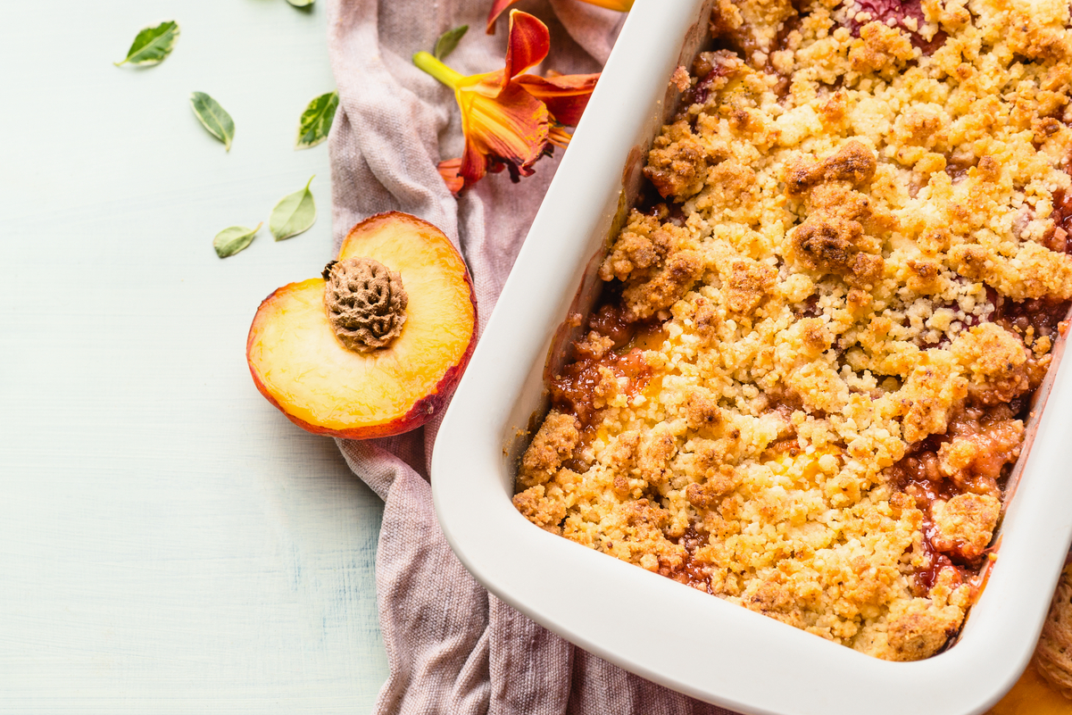 Weight Watchers Easy Peach Crumble in a white ceramic baking dish with a half of a peach and fresh tropical flowers next to it.