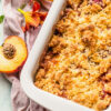 Closeup, overhead view of Weight Watchers Easy Peach Crumble in a white baking dish with a fresh, cut peach next to it.