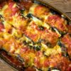 A close-up of a homemade Italian Eggplant Rollatini dish in a brown baking dish, featuring rolls of eggplant filled with cheese, covered in a chunky tomato sauce, and topped with melted golden cheese, all resting on a wooden surface.