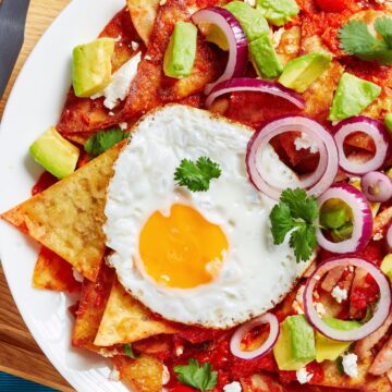 Close-up of a plate of Mexican chilaquiles with a fried egg on top, garnished with avocado slices, red onion rings, and cilantro, on a blue placemat.