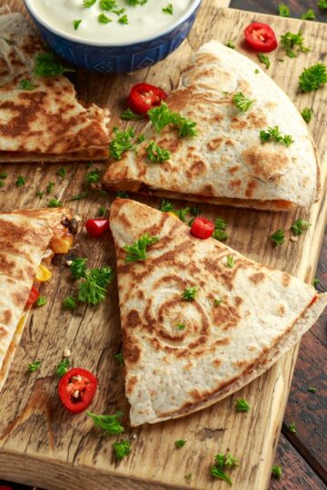 A close-up of a chicken and cheese quesadilla on a rustic wooden board, featuring melted cheese, with parsley and sliced red peppers for garnish.