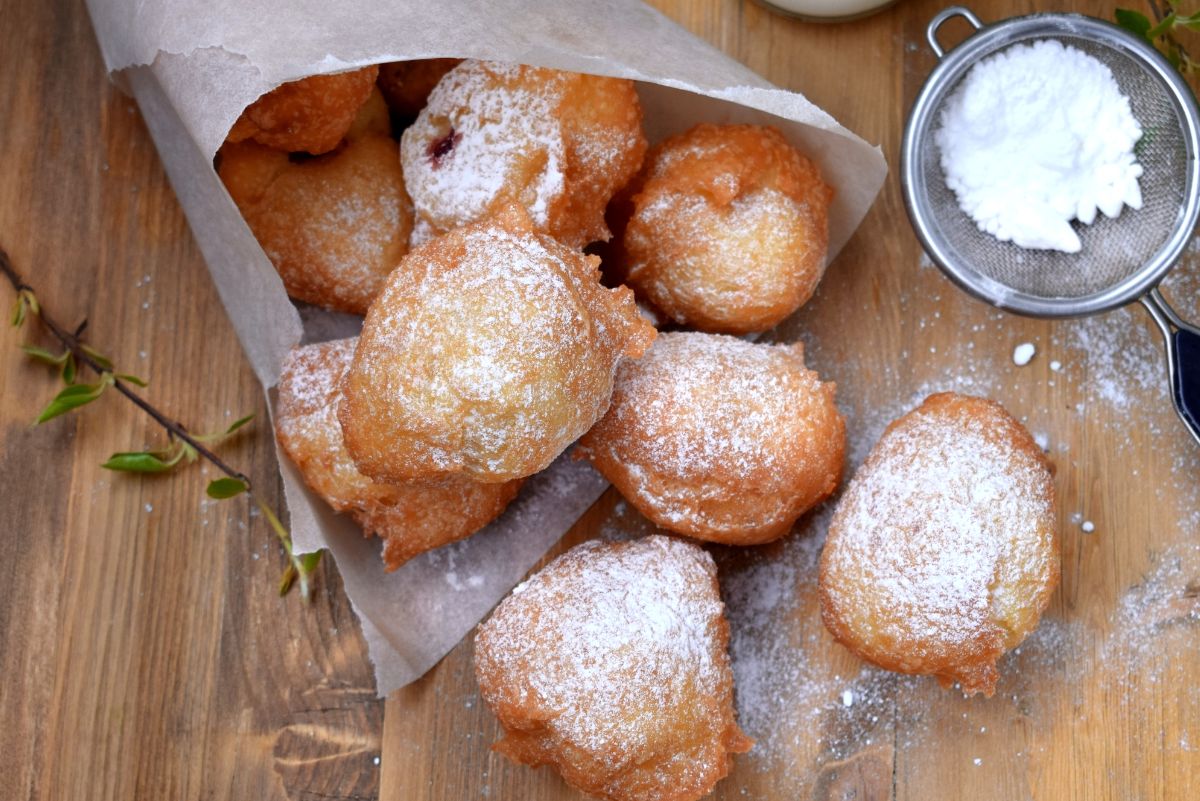A tempting batch of Weight Watchers-friendly beignets sit on a wooden surface, nestled in a paper-lined tray, suggesting they were cooked in an air fryer. These golden-brown pastries are generously dusted with powdered sugar, and one can spot a slight hint of a filling oozing out from one of the beignets. A fine sieve with more powdered sugar rests to the side, ready for additional dusting.