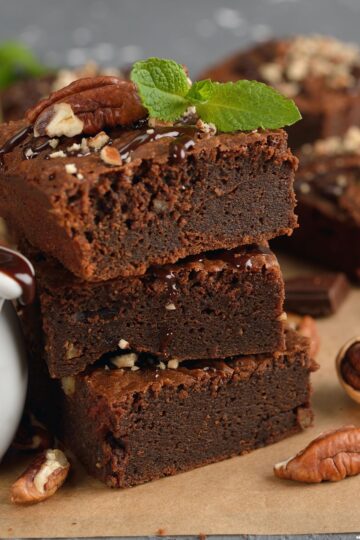 Close-up of a stack of lighter pecan brownies with a shiny chocolate drizzle, pecans, and a sprig of mint on top, emphasizing the moist texture and nutty addition.