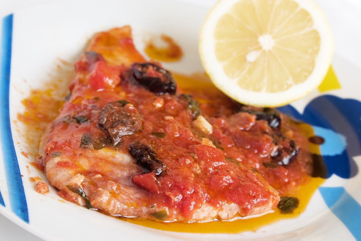 A plate of Quick and Easy Italian Fish Fillets, garnished with a lemon wedge, tomato sauce, and olives on a white plate with a blue and yellow striped border.