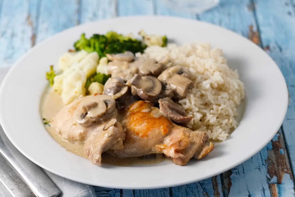 Crock Pot chicken rice casserole featuring tender browned chicken thighs smothered in creamy mushroom sauce with sliced mushrooms, served beside fluffy white rice and steamed broccoli and cauliflower on a white plate resting on a rustic blue wooden tabletop.