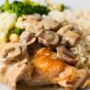 Crock Pot chicken rice casserole featuring tender browned chicken thighs smothered in creamy mushroom sauce, served over medium-grain rice with steamed broccoli and cauliflower on a white plate atop a blue wooden table.