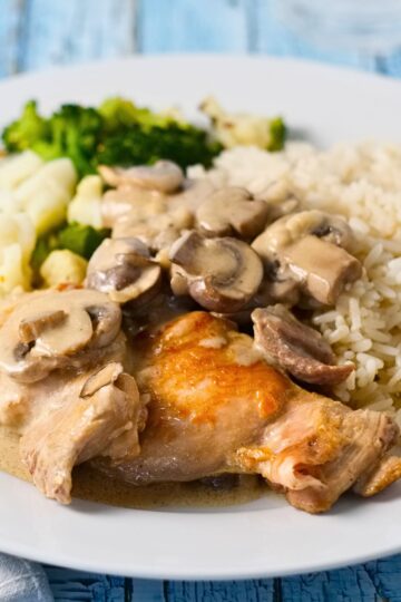Crock Pot chicken rice casserole featuring tender browned chicken thighs smothered in creamy mushroom sauce, served over medium-grain rice with steamed broccoli and cauliflower on a white plate atop a blue wooden table.