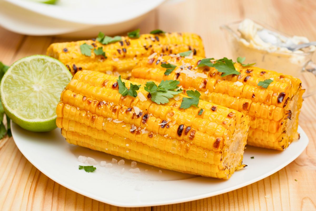 Air Fryer Roasted Corn. Charred golden corn on the cob garnished with fresh parsley and coarse sea salt, served on a white plate with a halved lime and a dish of creamy butter in the background.