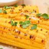 Air Fryer Roasted Corn. Golden roasted corn on the cob with light char marks, topped with fresh parsley and coarse sea salt, served on a white plate alongside a halved lime, with a dish of butter in the background.