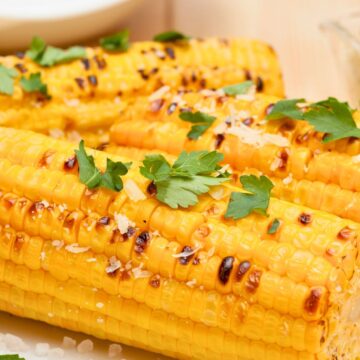 Air Fryer Roasted Corn. Golden roasted corn on the cob with light char marks, topped with fresh parsley and coarse sea salt, served on a white plate alongside a halved lime, with a dish of butter in the background.