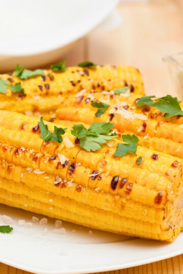 Air Fryer Roasted Corn. Golden roasted corn on the cob with light char marks, topped with fresh parsley and coarse sea salt, served on a white plate alongside a halved lime, with a dish of butter in the background.