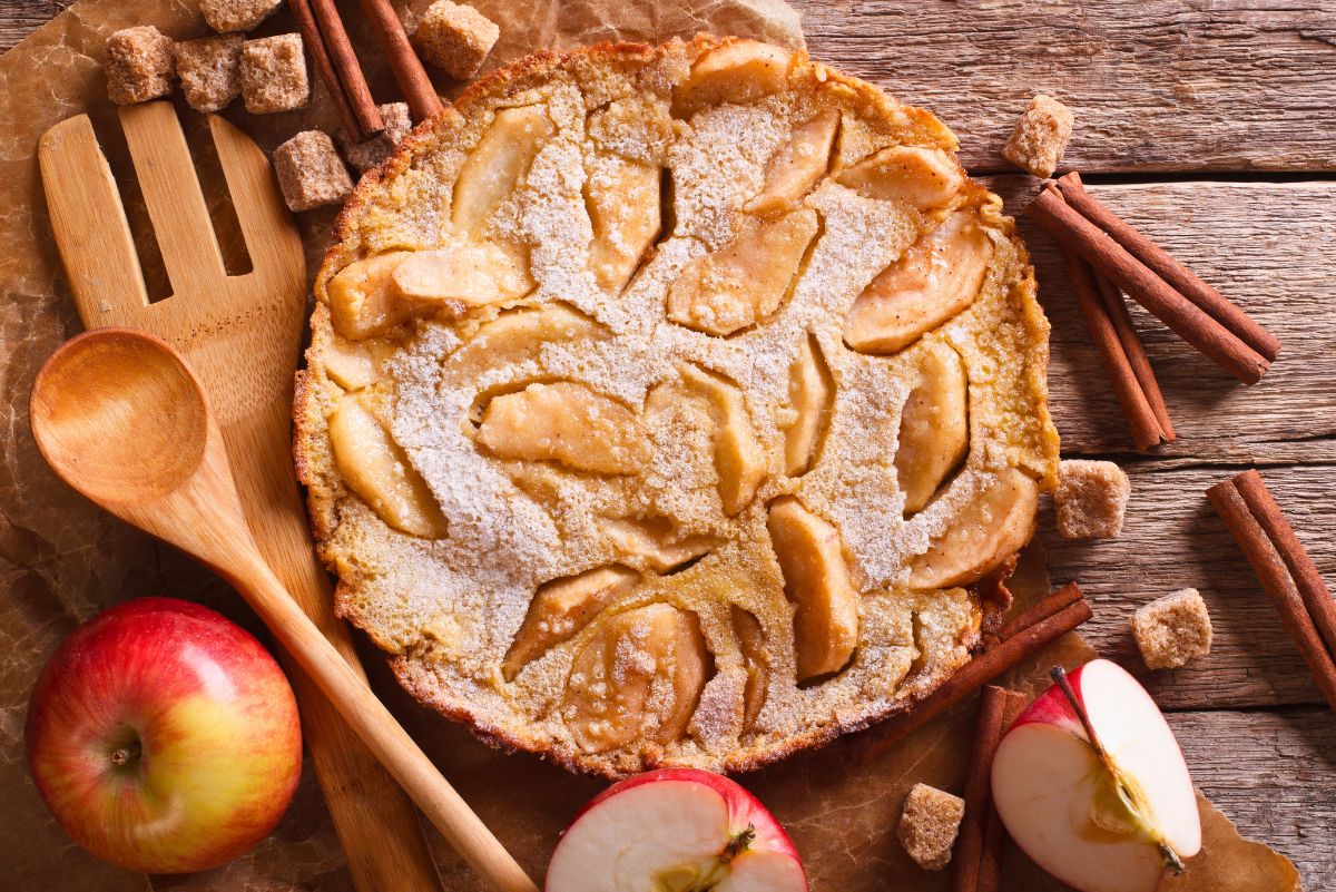 Golden baked apple cinnamon pancake topped with powdered sugar and layered with tender apple slices. Surrounded by fresh apples, cinnamon sticks, brown sugar cubes, and wooden cooking utensils on a rustic wooden table for a cozy fall breakfast scene.