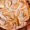 Close-up of a baked apple cinnamon pancake dusted with powdered sugar, featuring golden apple slices baked into the surface. Rustic background with cinnamon sticks, sugar cubes, and fresh apple halves adds a cozy fall kitchen feel.