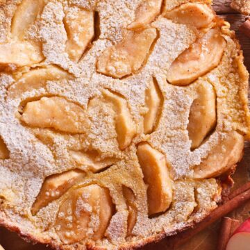 Close-up of a baked apple cinnamon pancake dusted with powdered sugar, featuring golden apple slices baked into the surface. Rustic background with cinnamon sticks, sugar cubes, and fresh apple halves adds a cozy fall kitchen feel.