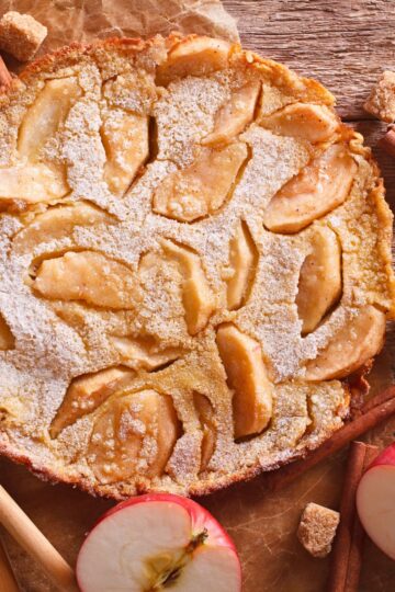 Close-up of a baked apple cinnamon pancake dusted with powdered sugar, featuring golden apple slices baked into the surface. Rustic background with cinnamon sticks, sugar cubes, and fresh apple halves adds a cozy fall kitchen feel.