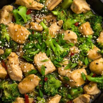 Close-up of Better-Than-Takeout Garlic Chicken and Broccoli stir fry in a skillet, with golden chicken pieces, fresh broccoli, sesame seeds, sliced scallions, and red chili peppers in a glossy garlic sauce.