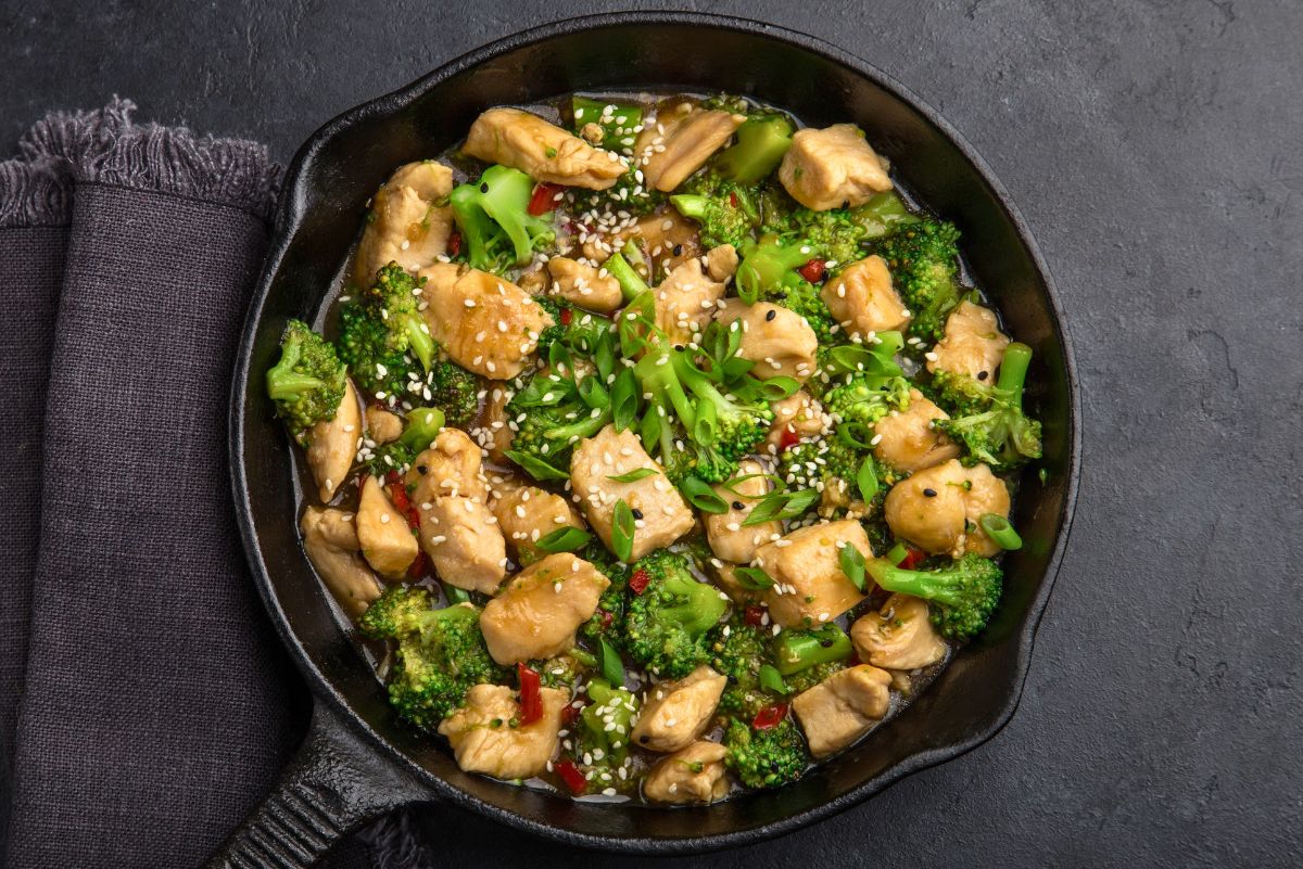 Overhead view of garlic chicken and broccoli stir fry in a cast iron skillet, featuring tender chicken chunks, green broccoli, scallions, sesame seeds, and red chili in a savory sauce on a dark background.