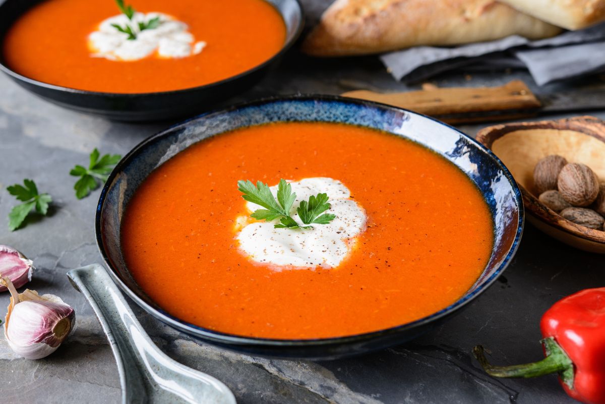 Two bowls of cozy roasted red pepper and pear soup with cream and parsley garnish, set on a stone surface with garlic, nutmeg, a red pepper, and crusty bread in the background.