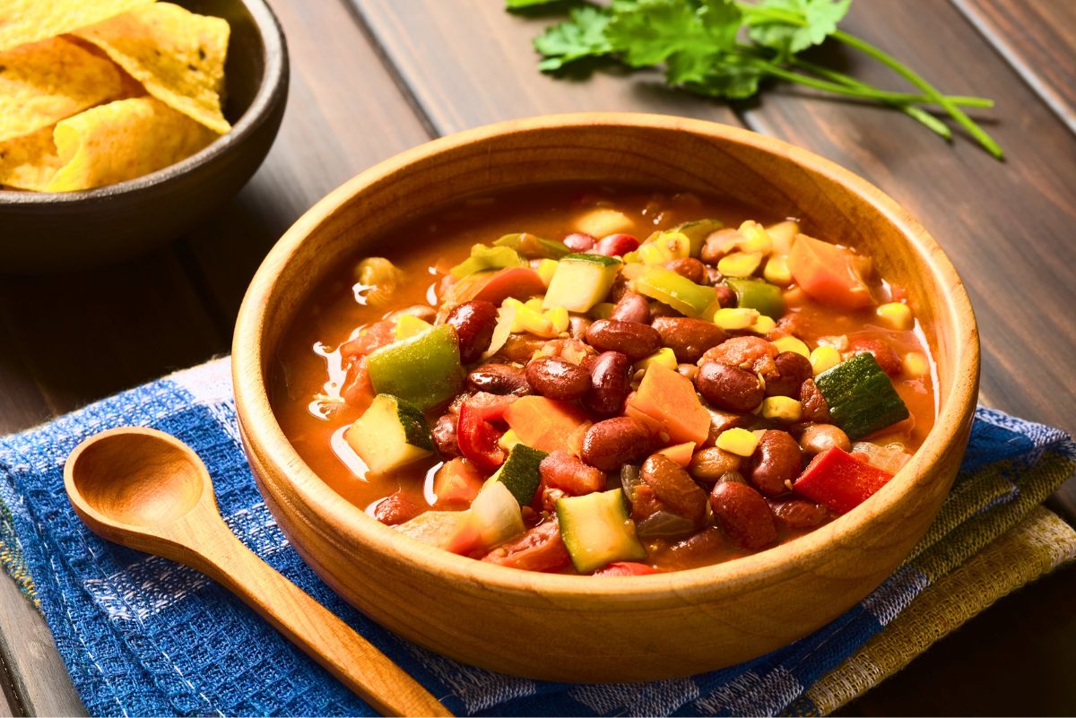 A rustic wooden bowl filled with Crockpot Spicy Vegetarian Chili, featuring kidney beans, corn, zucchini, bell peppers, carrots, and tomatoes in a rich broth. A wooden spoon rests on a blue cloth beside the bowl, with tortilla chips and fresh cilantro in the background. Perfect for a cozy, hearty plant-based dinner.
