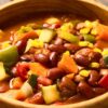 A close-up of a hearty bowl of spicy vegetarian chili filled with kidney beans, black beans, corn, zucchini, bell peppers, carrots, and tomatoes in a rich, savory broth. The vibrant mix of vegetables and beans sits in a rustic wooden bowl, showcasing a colorful and comforting plant-based meal.